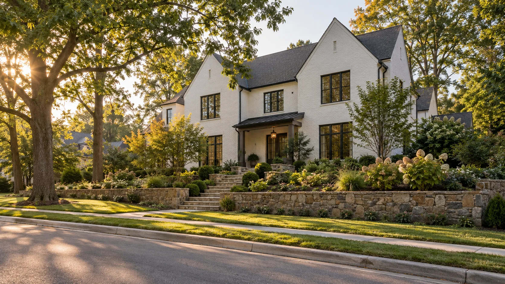 A white painted brick house with black windows seen from along the street, standing above a stone retaining wall with steps rising through layered planting to a covered entry in late afternoon light
