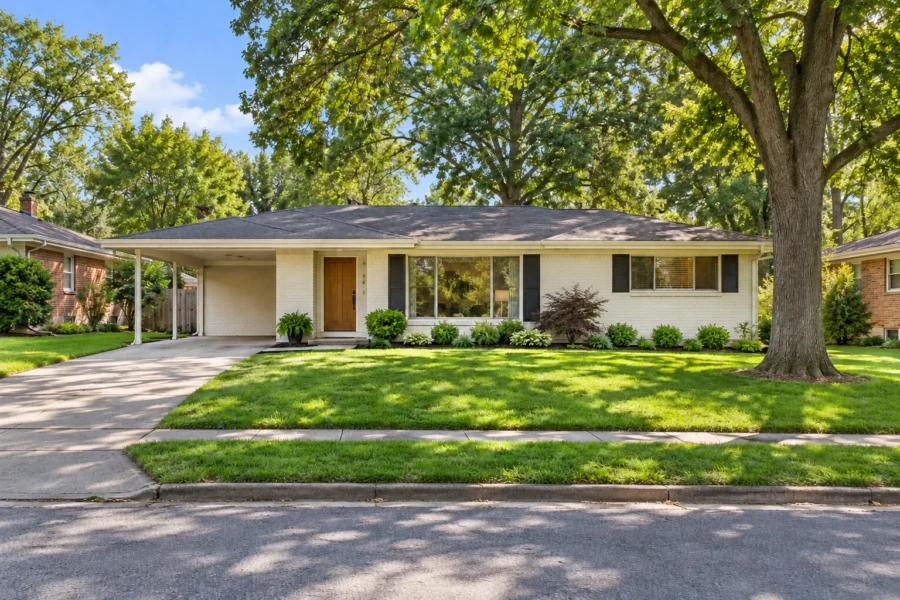 A single-storey mid-century ranch house in painted brick with a carport, mature shade trees and neighbouring houses on an established suburban street
