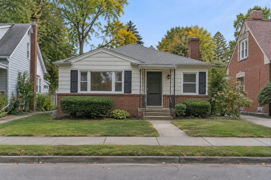 A modest older single storey house on a small lot, with brick and siding, clipped shrubs and a short concrete walk to the front steps