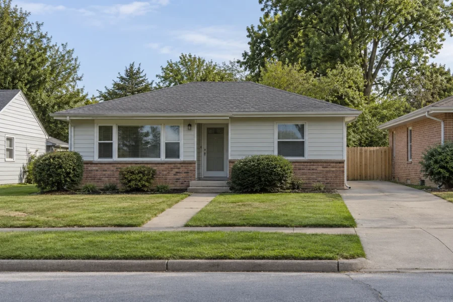 A modest starter scale single storey house in pale siding with a brick skirt, a small tidy lawn and clipped foundation shrubs