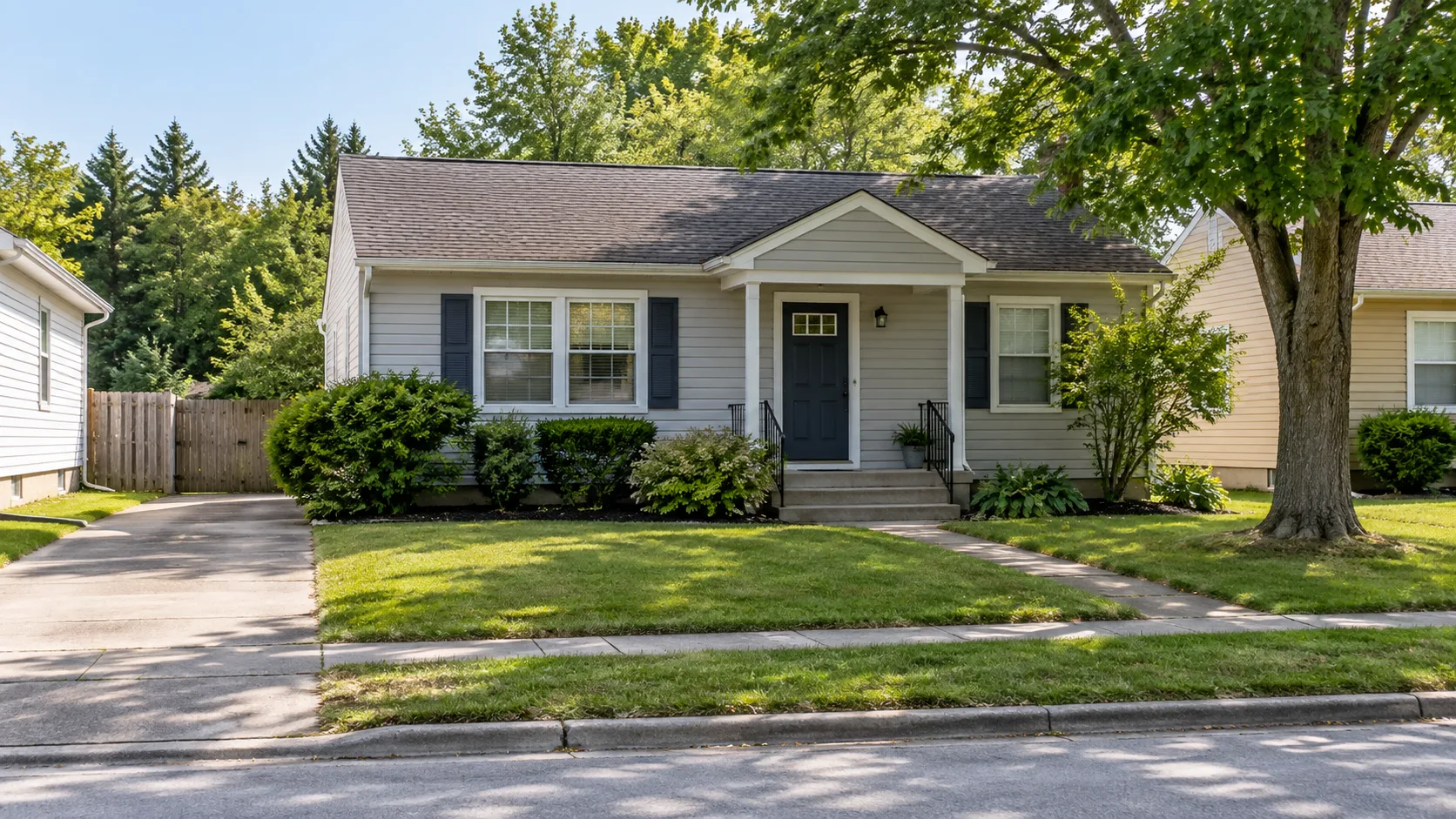 A small well kept single storey house with pale siding, a covered front stoop and a trimmed lawn on an ordinary residential street in summer daylight