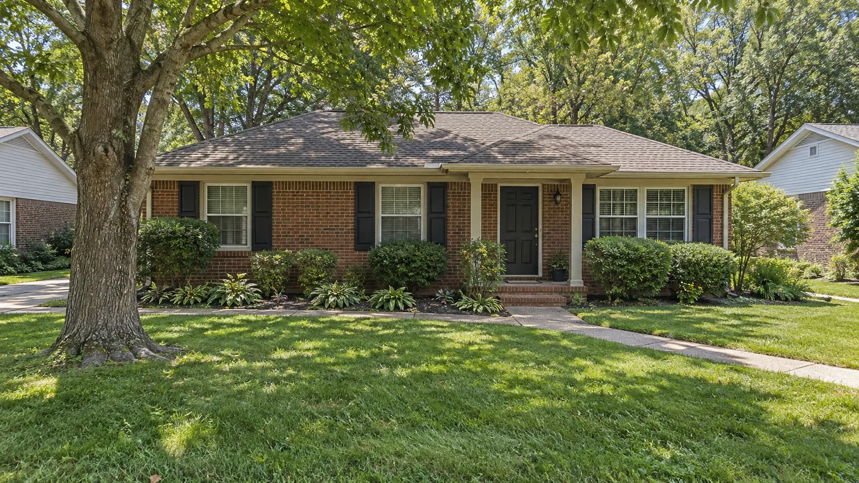 An ordinary single storey brick house in the American Southeast seen from the front yard under a large shade tree on a summer afternoon