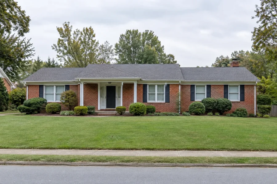 A plain single storey red brick ranch house with dark shutters and clipped foundation shrubs, seen straight on across a level lawn under flat overcast daylight