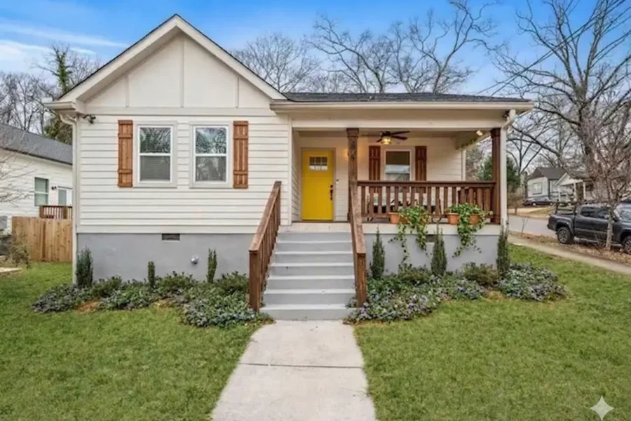 A white cottage with a yellow front door, a small porch and planted beds along the walk