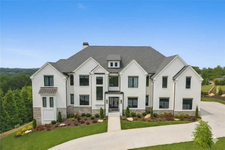 A large white two story house with a gabled roof and a curving driveway, with landscaped beds in front